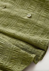 Oliivinvihreä kangas, jossa on teksturoitu, ryppyinen kuvio ja kaksi nappia. Materiaali on kevyt ja pehmeä koskettaa.