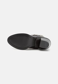Suela de zapato negra con patrón texturizado, fabricada en material de goma, forma redondeada y un ligero tacón. Incluye un detalle de tirante transparente.