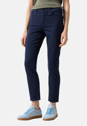 Personne portant un pantalon slim bleu marine et des baskets bleu clair avec des semelles en gomme, debout devant un fond uni.