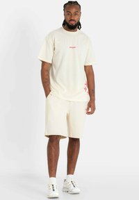 Beige oversized t-shirt with pink logo, paired with matching shorts. White sporty shoes with multi-colored accents. Simple, casual design.