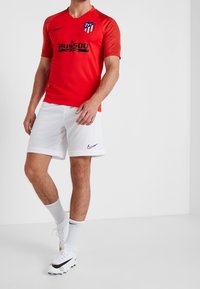 Hombre vestido con una camiseta de fútbol roja del Atlético de Madrid, pantalones cortos blancos de Nike, calcetines blancos y tacos blancos de Nike, posando contra un fondo blanco liso.