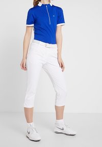 Niebieska koszulka polo z krótkim rękawem, białymi wykończeniami, z zamkiem przy kołnierzyku oraz białe spodnie capri. Model nosi białe buty sportowe.