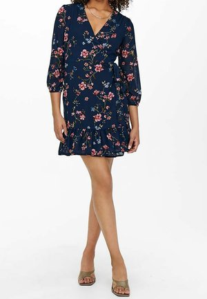 Kobieta ubrana w granatową sukienkę kopertową z różowym kwiatowym wzorem i falbaną na dole, w beżowych sandałach na obcasie z odkrytymi palcami, na białym tle.