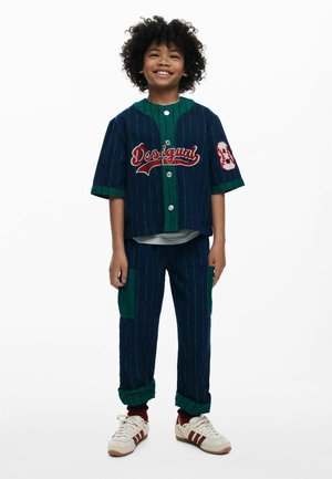 Bambino sorridente con capelli ricci che indossa una camicia a maniche lunghe a righe blu e verdi della "Desigual", pantaloni abbinati, calzini rossi e scarpe da ginnastica bianche.