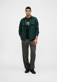 Zielona wełniana kurtka na czarnym t-shircie z grafiką, szare spodnie i czarne buty. Kurtka ma dwie kieszenie na piersi oraz zapięcie na guziki.