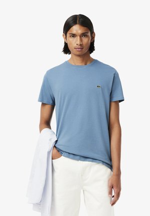 Light blue cotton T-shirt with a round neck and short sleeves. Features a small green Lacoste logo on the chest. Styled with white pants and a striped shirt.