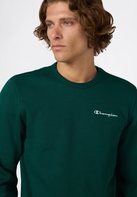 Sudadera verde de mangas largas con cuello redondo. Presenta un pequeño logo blanco de "Champion" en el lado izquierdo del pecho. Material de algodón suave.