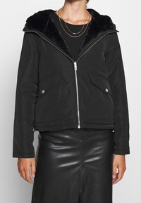 Kvinna som bär en svart jacka med dragkedja och pälskrage, svart topp, lager av guldhalsband och en svart läderkjol med framslits.