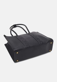 Sac cabas rectangulaire noir avec doubles poignées, clous dorés aux coins inférieurs et bandes verticales embossées du nom de la marque.