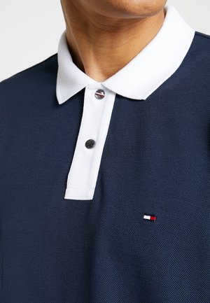 Mand iført navyblå poloshirt med hvid krave og knapstolpe, med to metal knapper og et lille rødt, hvidt og blåt logo på brystet.