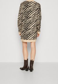 Personne portant une robe-pull imprimé zèbre beige et noir jusqu'aux genoux avec des manches longues et des bottines marron foncé à talons, posant devant un fond uni.