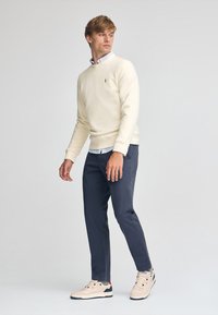 Felpa beige chiara, pantaloni blu navy e sneakers beige. Design casual con una texture morbida. Camicia bianca visibile al colletto.