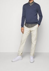 Pull en V marine porté sur une chemise à carreaux boutonnée, associé à un pantalon chino crème et des baskets montantes blanches, présenté en position debout.