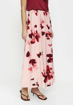 Žena oblečená v svetloružovej midi sukni s červenými kvetinovými vzormi, v kombinácii so červeným tielkom bez rukávov a hnedými sandálmi na opätku.