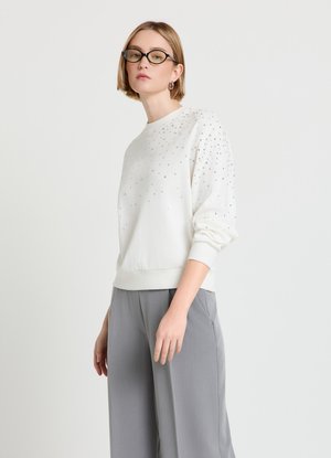Jeune femme portant des lunettes noires, un sweat-shirt blanc avec de petits clous argentés, et un pantalon gris ample, debout devant un fond clair uni.