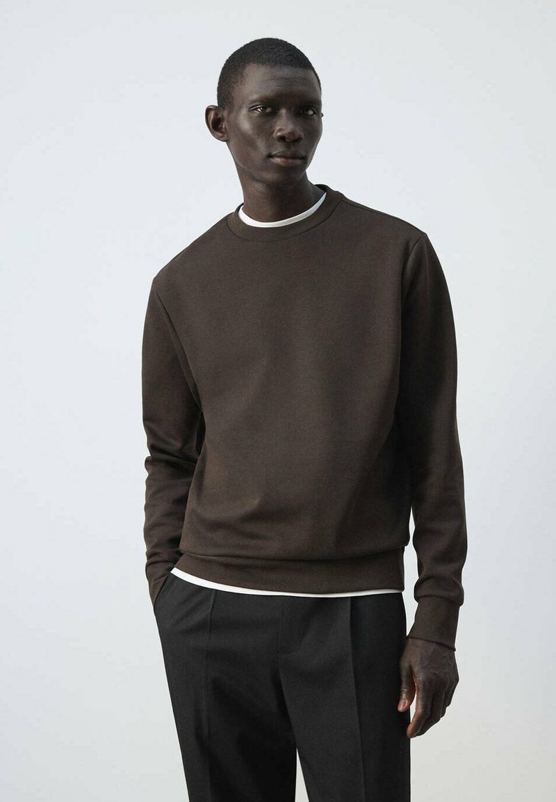 Homme à la peau foncée portant un sweat-shirt marron foncé sur une chemise blanche et un pantalon noir, debout devant un fond uni gris clair.