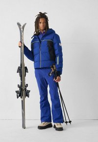 Person in blauer Skibekleidung hält schwarze Skier in einer Hand und Skistöcke in der anderen, steht vor weißem Hintergrund.