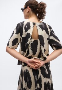 Μπλούζα με κοντό μανίκι σε μπεζ με μαύρο αφηρημένο σχέδιο· διαθέτει άνοιγμα στην πλάτη και απαλή υφή. Συνδυάζεται με φούστα σε συντονισμένο χρώμα.