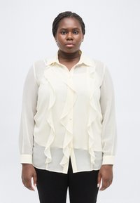 Γυναίκα με πλεξούδες, φορούσα διαφανές κρεμ πουκάμισο με βολάν και μαύρο παντελόνι, στέκεται μπροστά σε λευκό, απλό φόντο.