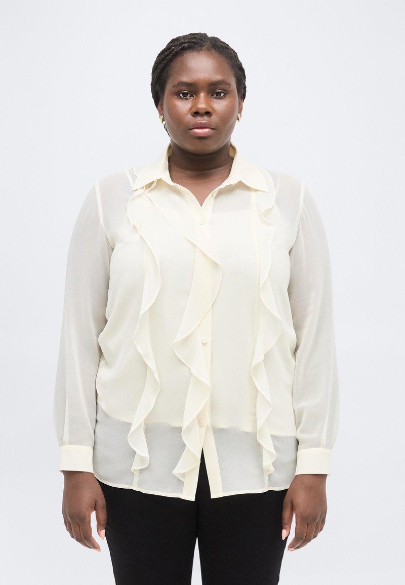 Γυναίκα με πλεξούδες, φορούσα διαφανές κρεμ πουκάμισο με βολάν και μαύρο παντελόνι, στέκεται μπροστά σε λευκό, απλό φόντο.