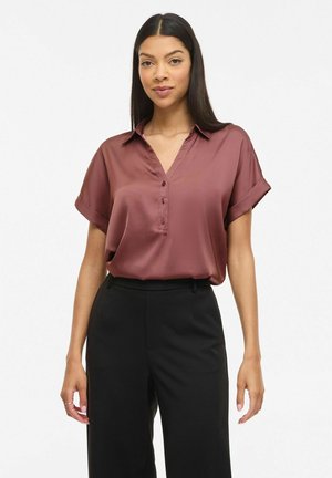 Femme aux longs cheveux foncés portant un chemisier mauve à manches courtes boutonné, rentré dans un pantalon noir taille haute, debout devant un fond blanc.