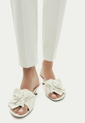 Füße in weißen, niedrigen Slip-On-Sandalen mit großen Stoffschleifen mit Perlenakzenten, kombiniert mit weißen kurzen Hosen.