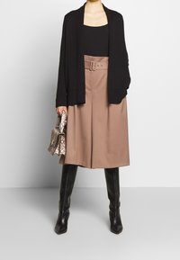 Svart öppet cardigan, svart linne, beige hög midja vida byxor med bälte, knähöga svarta stövlar och en mönstrad handväska.