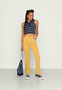 Top cropped azul listrado com colarinho, combinado com calças amarelas de cintura alta e bocados largas. Modelo segura uma mala azul, usando sapatilhas pretas.