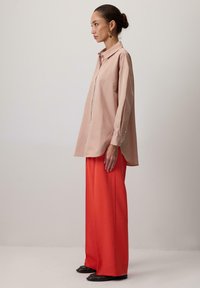 Gaiši rozā pārāk liels krekls ar apkakli, pogu priekšu un garām piedurknēm; apvienots ar spilgti oranžām plašām biksēm; gluda tekstūra.