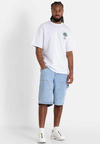 Wit T-shirt met een groen en rood palmboomafbeelding, gecombineerd met lichtblauwe denim shorts met zijzakken. Witte sneakers maken de look compleet.