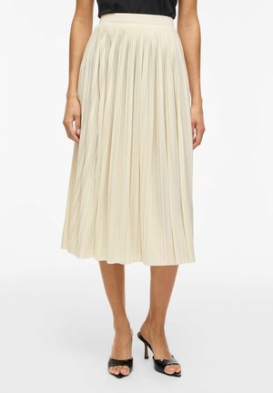 Femme portant une jupe midi plissée couleur crème avec un haut noir et des chaussures noires à talons ouverts, debout sur un fond blanc.