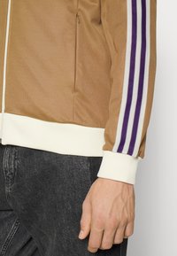 Brun zip-up jacka med en krämfärgad ribbad nederkant, prydd med lila och vita ränder längs ärmarna. Materialet verkar slätt och strukturerat.