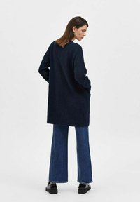 Cardigan lungo in maglia blu navy con vestibilità ampia, dotato di scollo rotondo e spalle scese. Abbinato a jeans a gamba larga e scarpe nere.