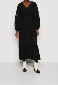 Abito nero a maniche lunghe con pannelli in pizzo, scollatura a V e chiusura frontale con bottoni. Presenta una gonna a strati e un tessuto testurizzato. Indossato con stivali bicolore.