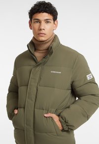 Olivenfarvet pufferjakke med struktureret finish, høj krave og to sidelommer. Har et "Guess Jeans" logo på brystet og på ærmet.