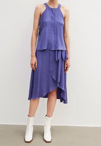 Purple dress with a textured wave pattern, layered design, asymmetrical hem, and a tied detail. Paired with white ankle boots.