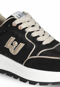 Baskets noires avec une finition brillante, semelle intercalaire blanche, lacets beiges et logo avec accents dorés. Présente une tige texturée et une semelle extérieure en caoutchouc.