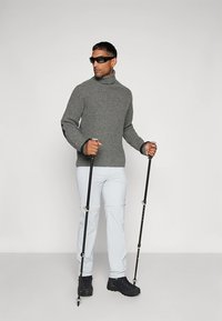 Šedý žebrovaný rolák, světle modré kalhoty, černé turistické boty a trekové hole. Model má na sobě sluneční brýle a stojí na neutrálním pozadí.