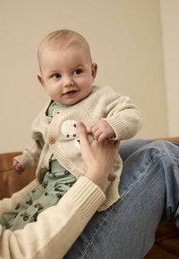 Cardigan en tricot beige clair avec des boutons sur le devant et un motif de mouton, porté par-dessus une combinaison verte à motifs. Texture douce et coupe décontractée.