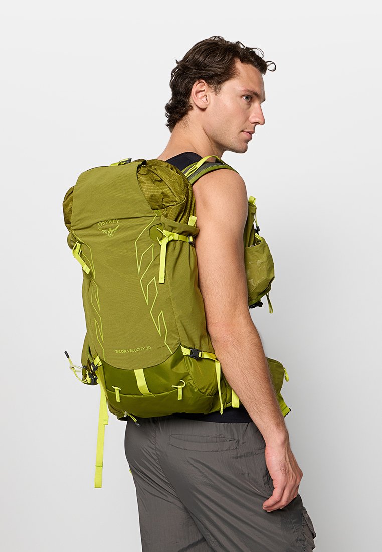 Mochila verde de nailon con múltiples compartimentos, correas ajustables y adornos de color amarillo brillante. Presenta un diseño aerodinámico y elegante.