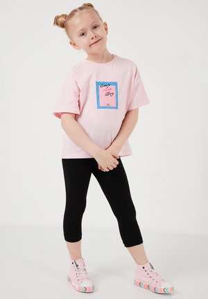 Ung pige med blondt hår i knolde iført en lyserød T-shirt, sorte leggings og lyserøde sneakers, stående med hænderne foldet.