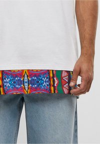 Partea inferioară a corpului unei persoane îmbrăcate cu blugi de culoare albastru deschis și o cămașă albă cu un tiv colorat și cu modele vizibile dedesubt.