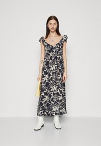 Robe maxi avec un motif floral bleu marine, manches volantées, décolleté en V et taille cintrée, accompagnée de bottines blanches et d'un sac à main jaune.
