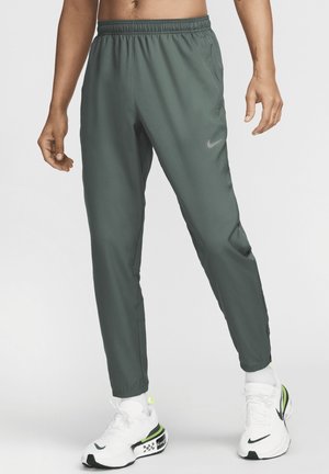 Mann trägt dunkelgrüne Nike-Trainingshose und weiße Nike-Sneaker mit neon-grünen Akzenten, steht vor schlichtem Hintergrund.