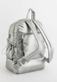 Srebrny plecak wypełniony poduszką o teksturowanym metalicznym wykończeniu, z regulowanymi paskami, przednią kieszenią na zamek błyskawiczny oraz dekoracyjnym sznurem do ciągnięcia przy zamku.