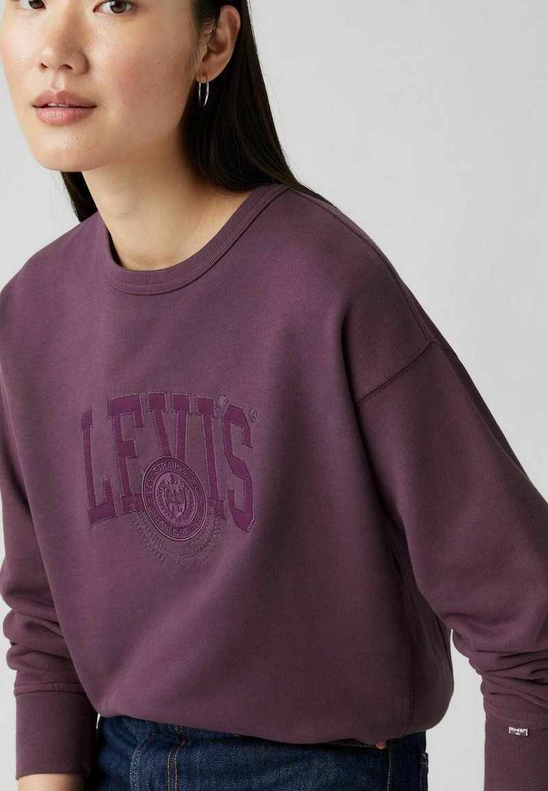 Lila Sweatshirt mit einem großen, gestickten "LEVI'S"-Logo und einem kreisförmigen Design. Hergestellt aus weichem, strukturiertem Stoff mit gerippten Bündchen.