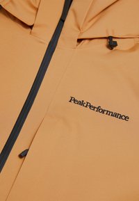 Giacca impermeabile arancione con una texture liscia, caratterizzata da una zip nera e dal logo "Peak Performance" ricamato in nero.