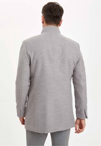 Grau maßgeschneiderte Jacke mit hohem Kragen, glatter Textur, einem einzelnen Rückenschlitz und langen Ärmeln, getragen über grauen Hosen.