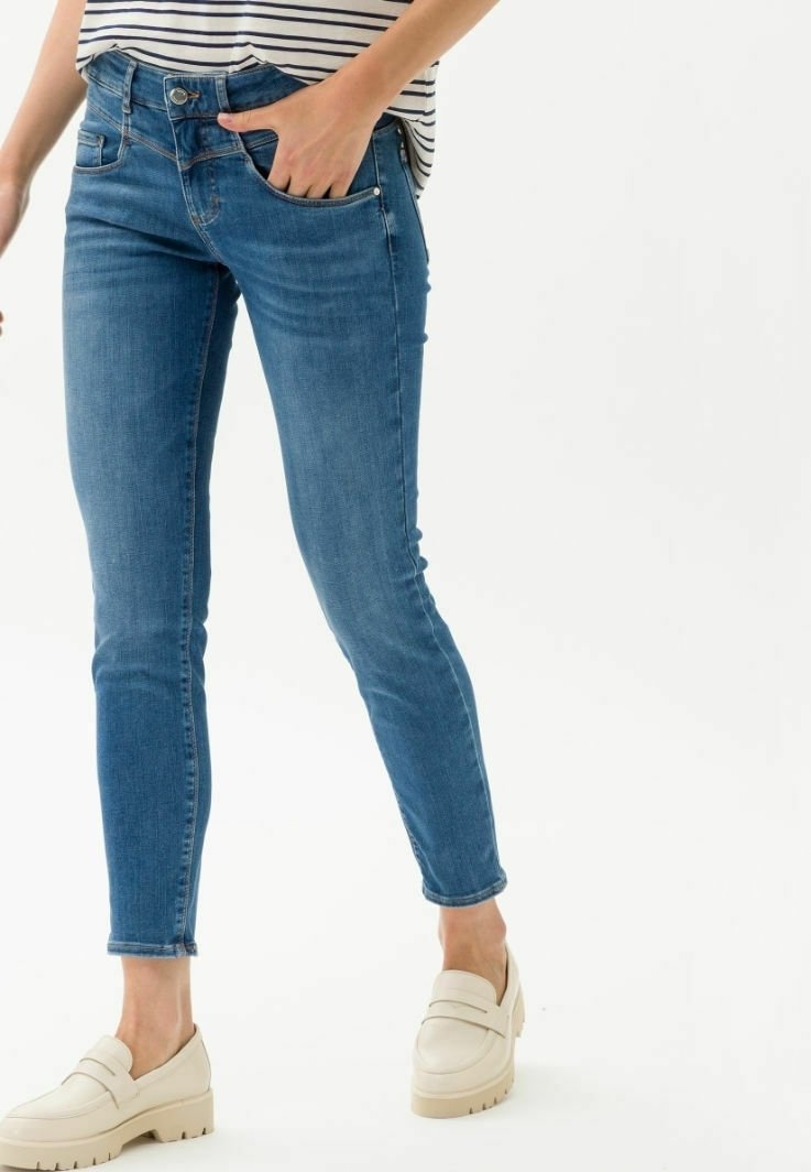 Femme portant un jean skinny bleu et des mocassins compensés crème, une main dans la poche avant, sur fond blanc.