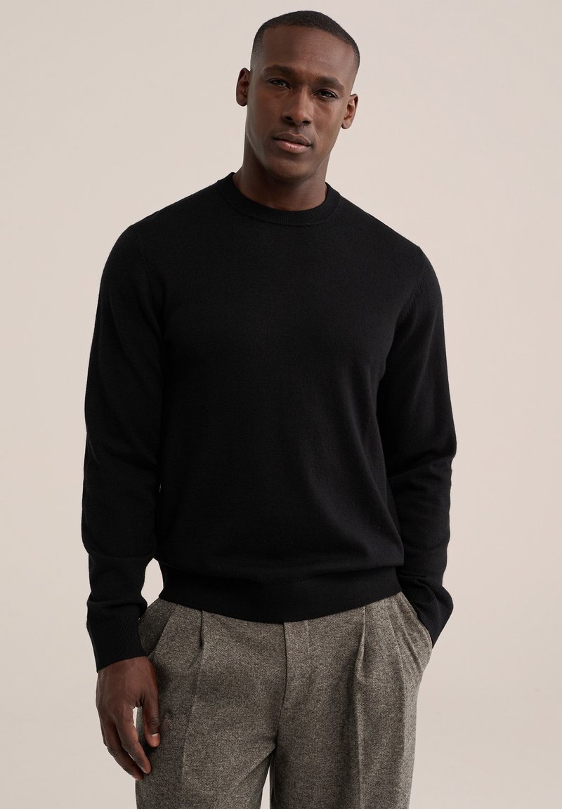 Homme portant un pull noir à manches longues et un pantalon plissé gris, les mains dans les poches, devant un fond beige uni.
